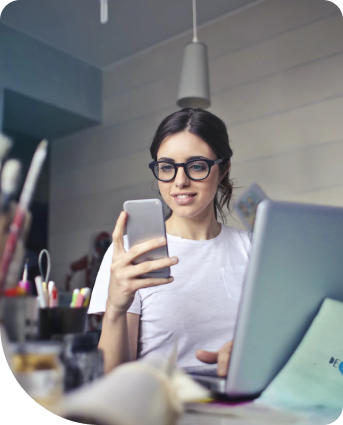 Mulher usando óculos sentada em frente ao notebook utilizando um celuar